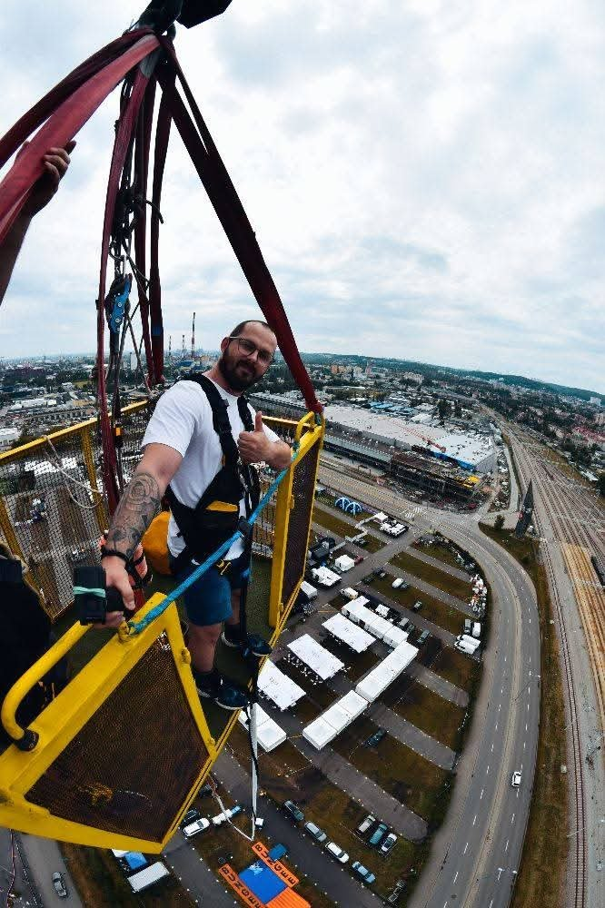 Sebastian w uprzęży na platformie wysoko nad miastem, z kciukiem w górę