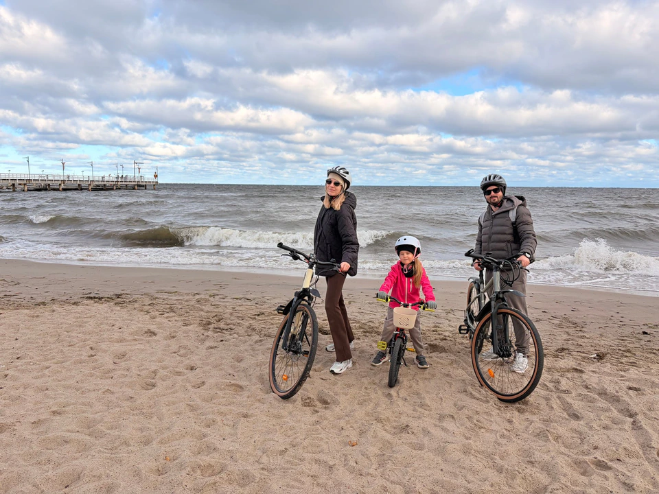 Sebastian z rodziną na rowerach na plaży, za nimi morze i molo